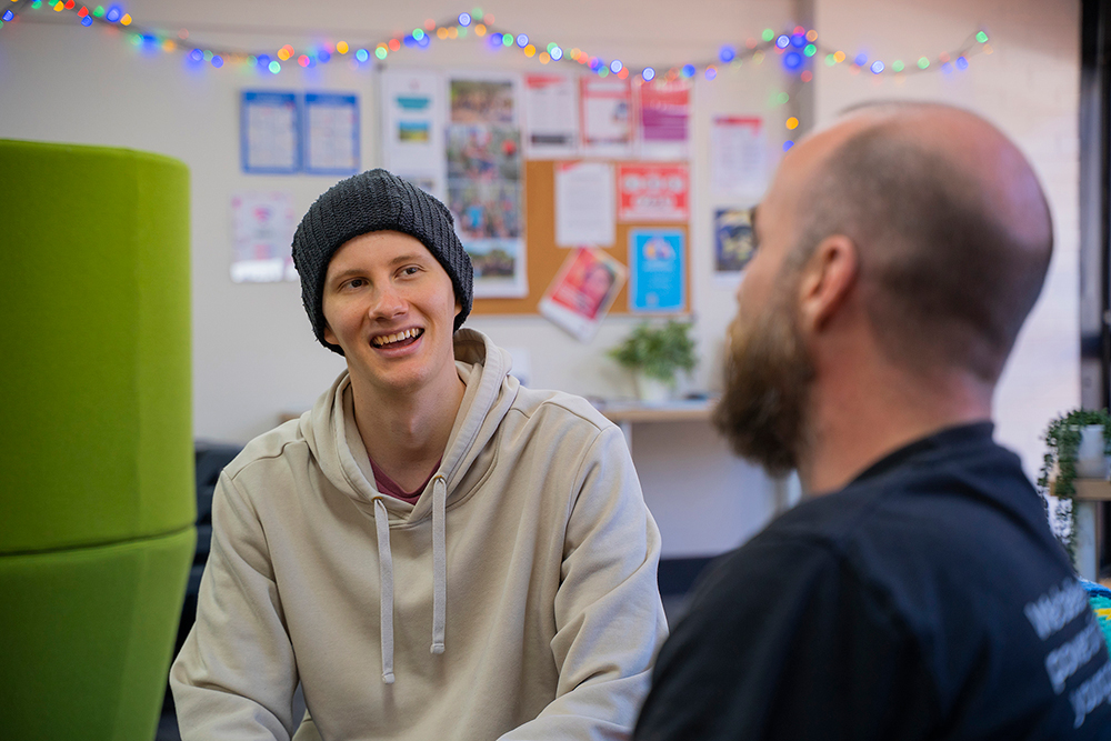 A smiling young person in a beanie chatting with a YMCA staff member in a cosy youth centre room decorated with fairy lights and a noticeboard