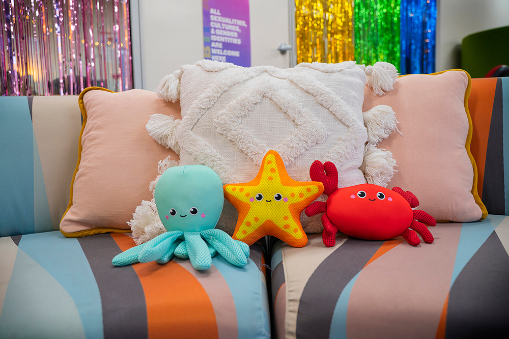 Three cute ocean-themed plush toys (an octopus, starfish, and crab) sitting on a colourful sofa with cushions, in front of rainbow tinsel curtains and an inclusivity sign