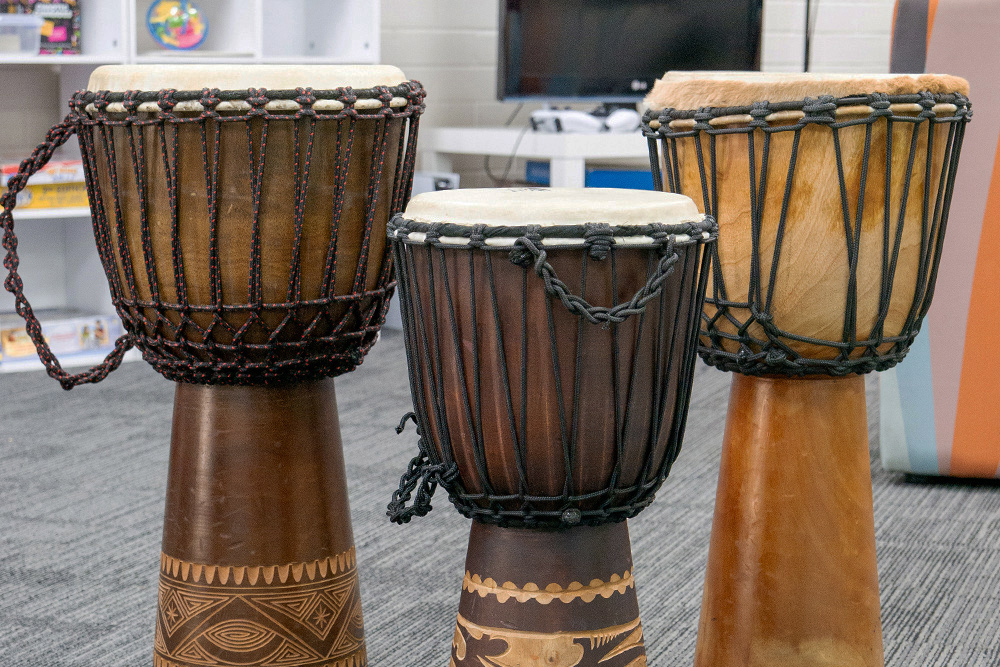 Three djembe drums with carved wooden bases displayed on the floor of a youth centre room