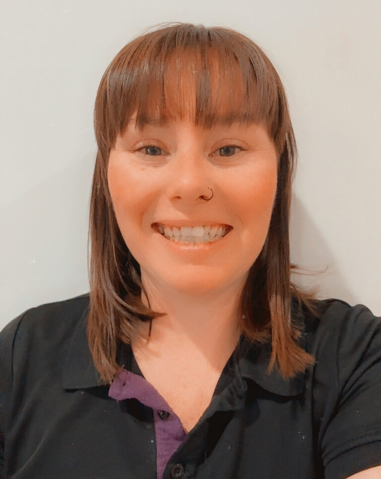 Smiling OSHC educator in YMCA uniform with name badge standing in outdoor garden setting