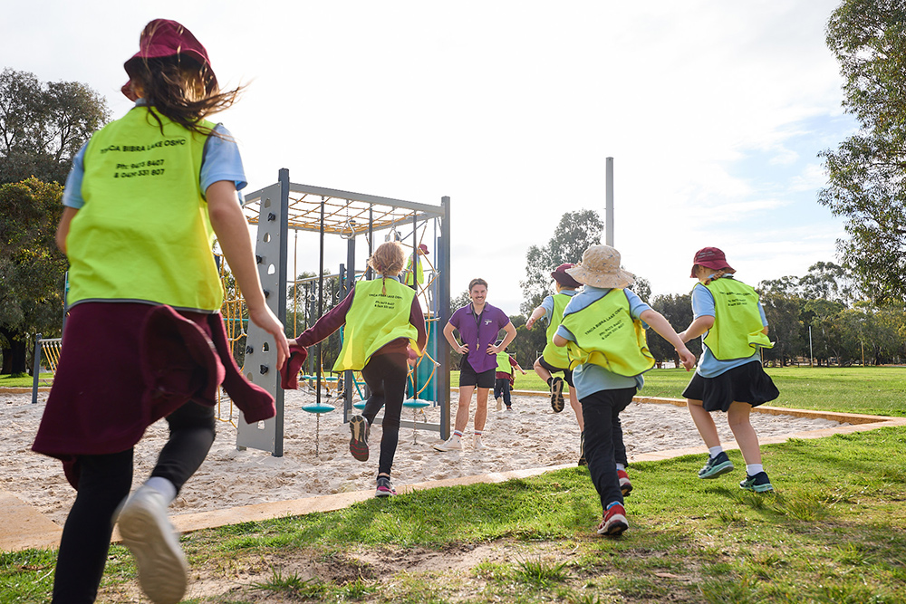 Children in Y WA hi-vis vests running towards playground equipment supervised by a staff member outdoors.