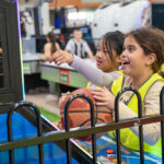 Two excited children in hi-vis vests playing an arcade basketball game during a Y WA OSHC excursion.