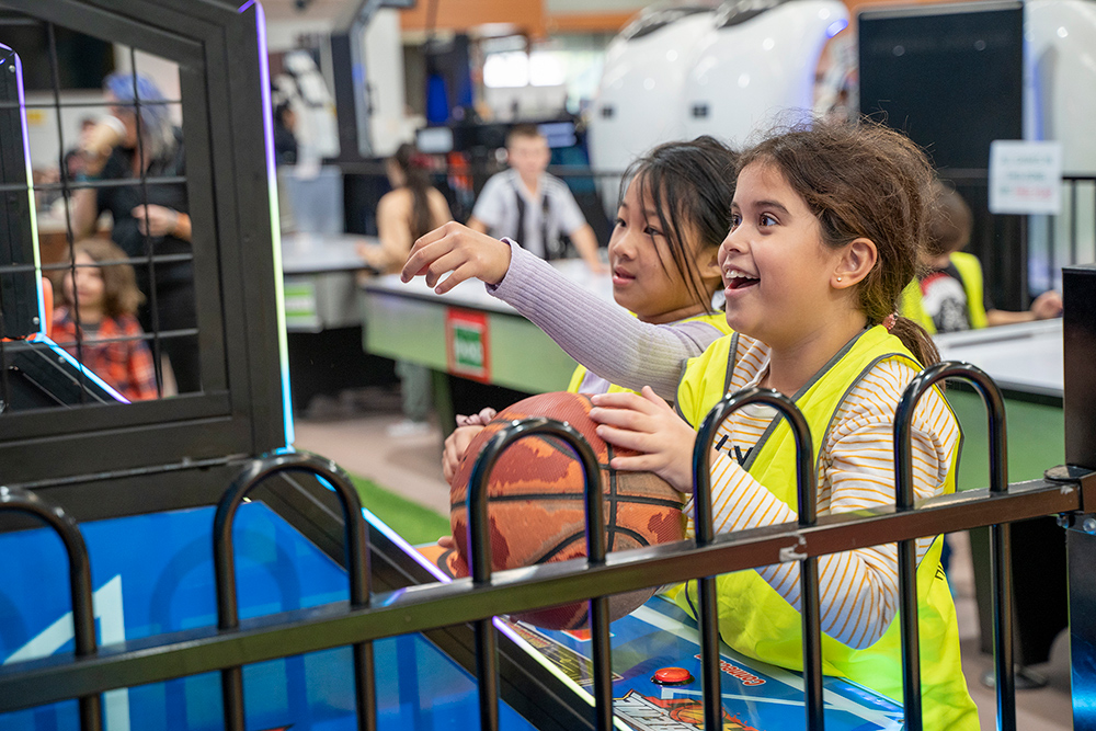 Two excited children in hi-vis vests playing an arcade basketball game during a Y WA OSHC excursion.