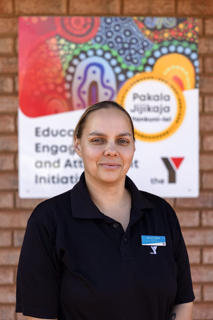 Sammi Ryder, Y WA program Pakala Jijikaja Yankuni-La Coordinator standing in front of a colourful program sign on a brick wall
