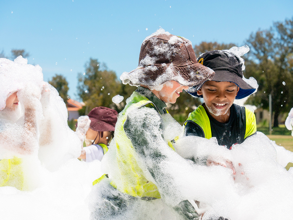Children in sun-safe hats enjoying outdoor foam play activity on sunny day at childcare