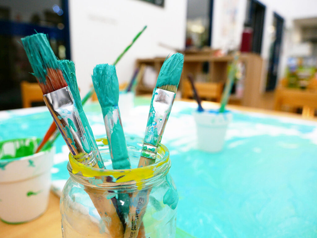 A glass jar holding several teal-painted brushes on an art table in an early learning centre classroom