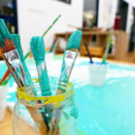 A glass jar holding several teal-painted brushes on an art table in an early learning centre classroom
