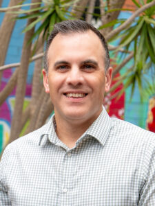 Smiling Y WA Board member James in a grey checked shirt, photographed outdoors against a colourful mural backdrop.