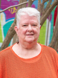 Smiling YMCA WA Board member Margaret in an orange top, photographed outdoors against a colourful mural backdrop.