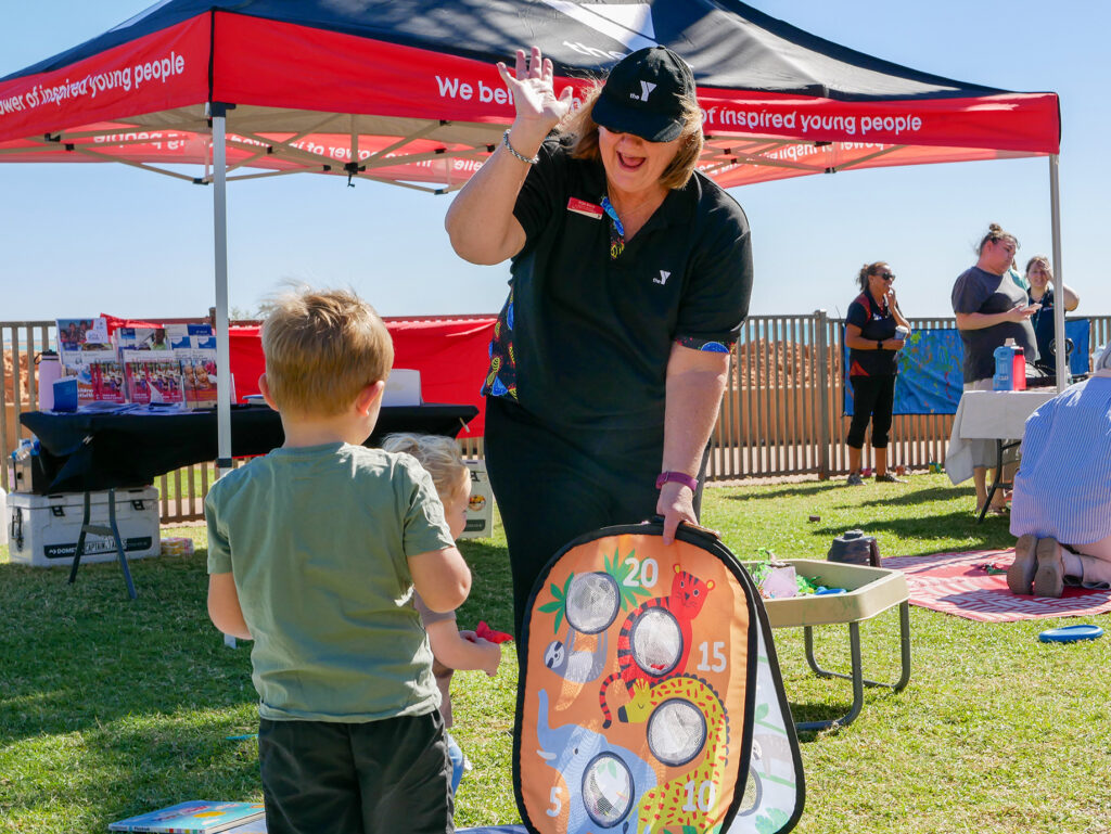 A Y WA staff member enthusiastically holds up a colourful bean bag toss game for a young child at an outdoor community event, with a Y WA marquee in the background