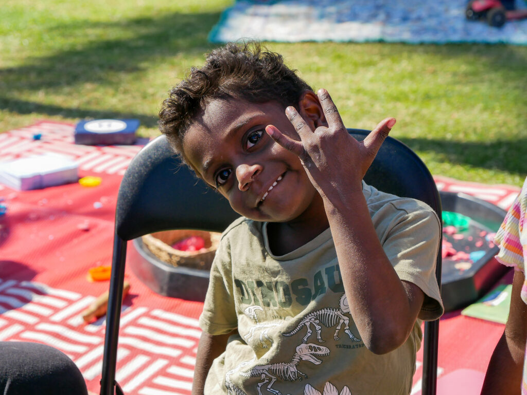 A young child in a dinosaur t-shirt waves and grins at the camera while sitting at an outdoor play activity table