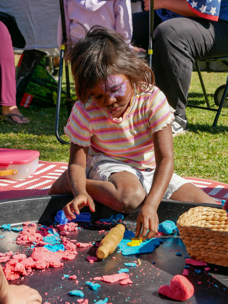A young girl with butterfly face paint sits on a mat and plays with pink and blue playdough and cutters at an outdoor event