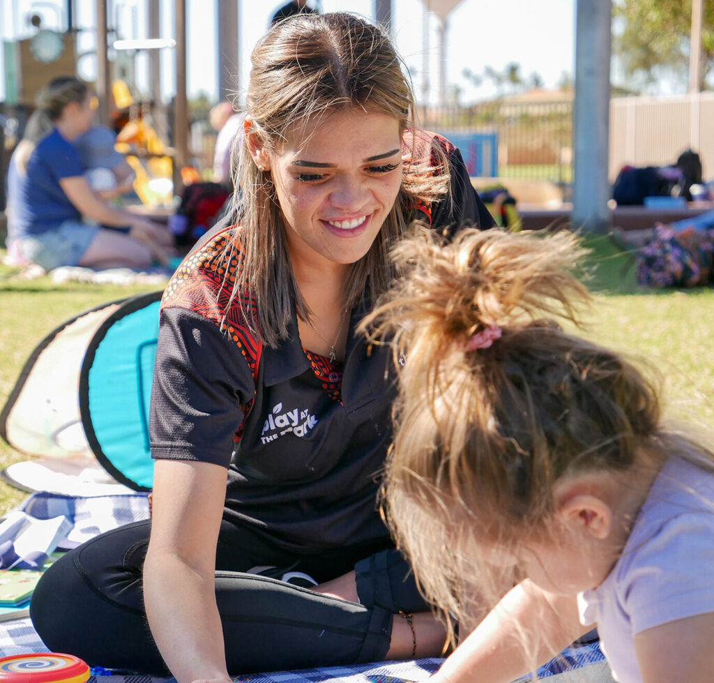 A Y WA worker in uniform smiles as she sits on a rug with a toddler exploring a busy board at an outdoor community event
