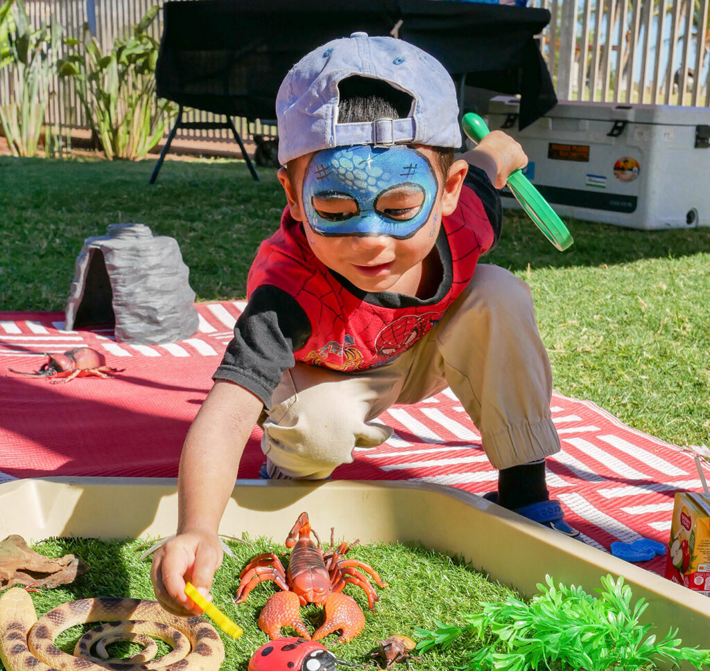 A young child with face paint and a cap leans over a sensory tray filled with plastic insects and reptile toys at an outdoor play event