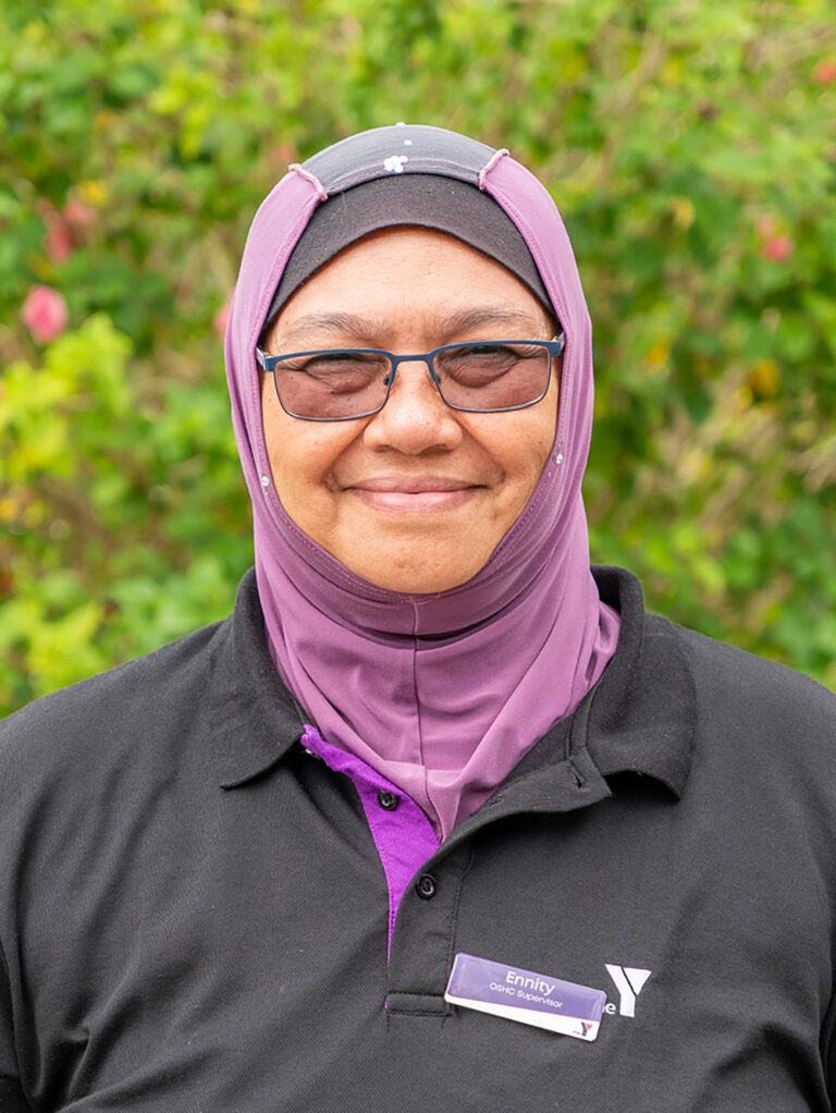 Smiling OSHC educator in YMCA uniform with name badge standing in outdoor garden setting