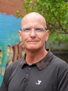 A smiling Y WA executive in a dark Y polo shirt, photographed outdoors against a colourful mural backdrop.