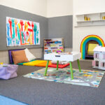 OSHC play area with rainbow cushions, dollhouse, children's handprint art and activity storage