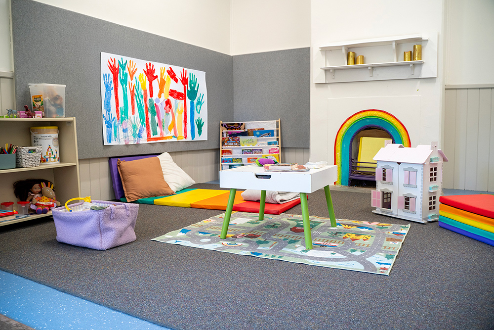 OSHC play area with rainbow cushions, dollhouse, children's handprint art and activity storage