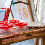 Paint containers and brushes arranged on splattered easel at OSHC creative arts station
