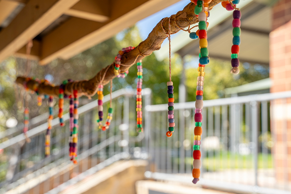 Colorful beaded craft hanging from tree branch at OSHC outdoor play area