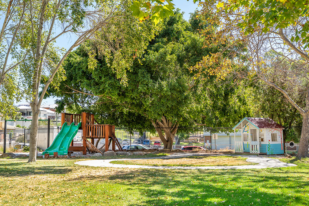 OSHC outdoor play area with wooden playground equipment, cubby house and shade trees