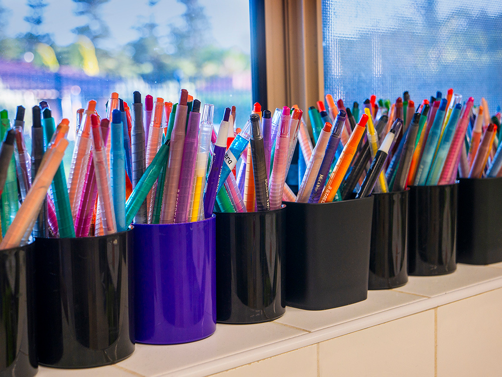 Row of pencil holders filled with colorful markers and pens at OSHC art station