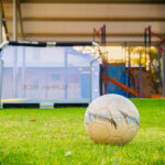 Soccer ball on grass field with goal post and playground equipment at OSHC outdoor area
