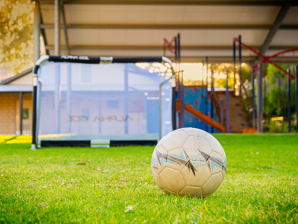 Soccer ball on grass field with goal post and playground equipment at OSHC outdoor area