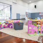 Colourful plastic dollhouse and treehouse play sets on rug in spacious OSHC hall