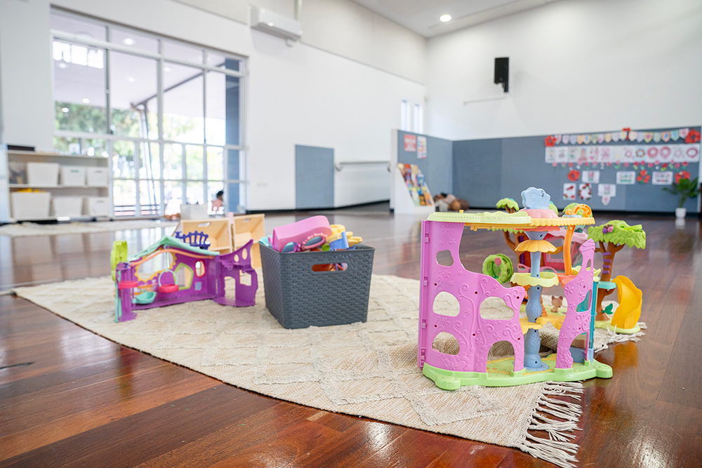 Colourful plastic dollhouse and treehouse play sets on rug in spacious OSHC hall