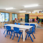 Clean OSHC room with white tables, blue chairs, bag hooks and children's decorations