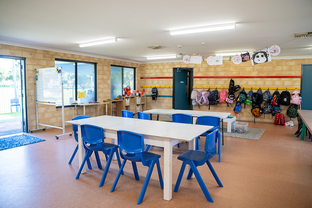 Clean OSHC room with white tables, blue chairs, bag hooks and children's decorations