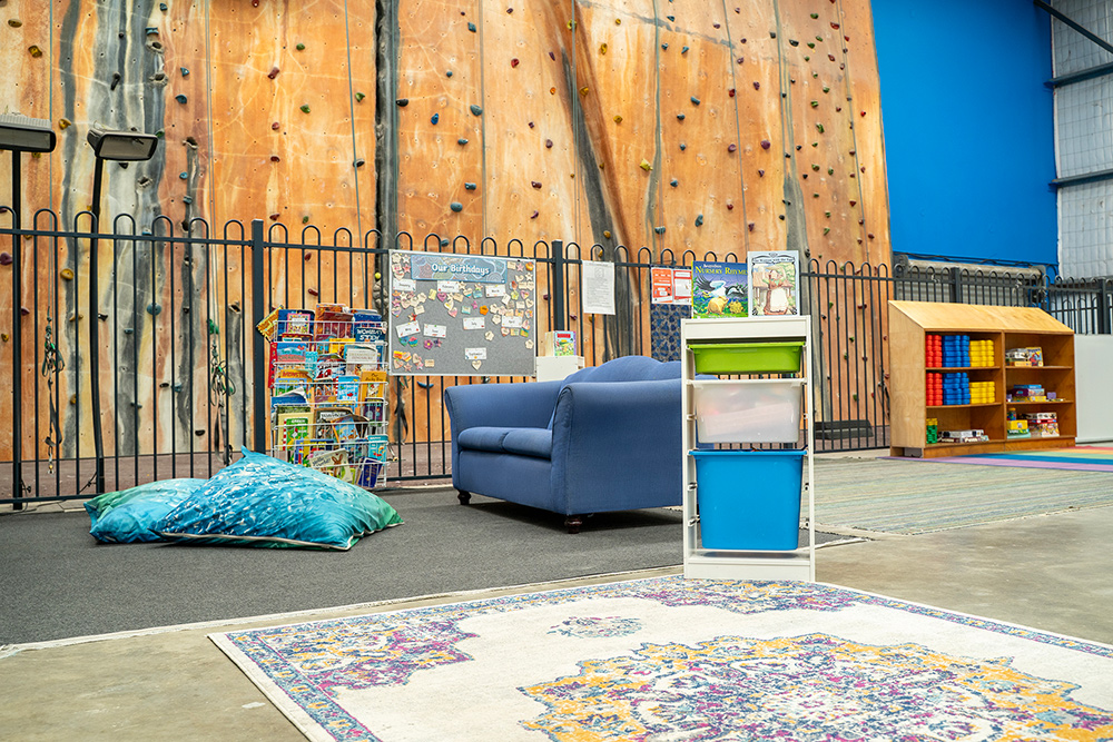 Relaxed OSHC reading nook with blue sofa, floor cushions, patterned rug, and indoor rock climbing wall backdrop.