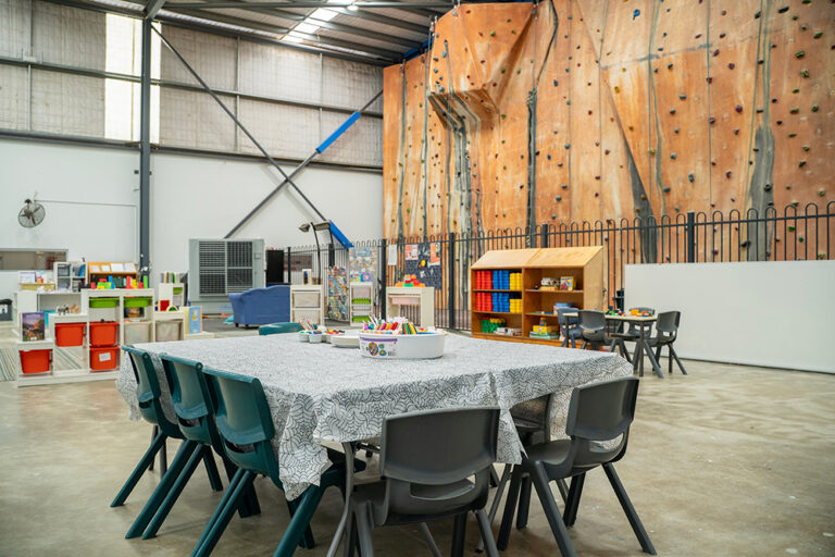 Unique OSHC space with activity tables, colourful storage, and a large indoor rock climbing wall backdrop.