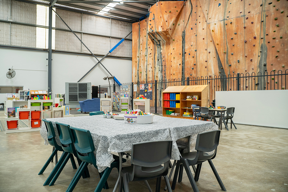 Unique OSHC space with activity tables, colourful storage, and a large indoor rock climbing wall backdrop.