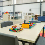 Colourful LEGO constructions on a table in a spacious OSHC activity room with storage shelving and seating areas.