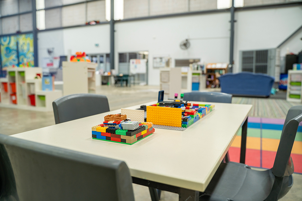 Colourful LEGO constructions on a table in a spacious OSHC activity room with storage shelving and seating areas.