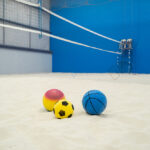 Colourful sports balls on a sand court inside an indoor sports facility with volleyball net.