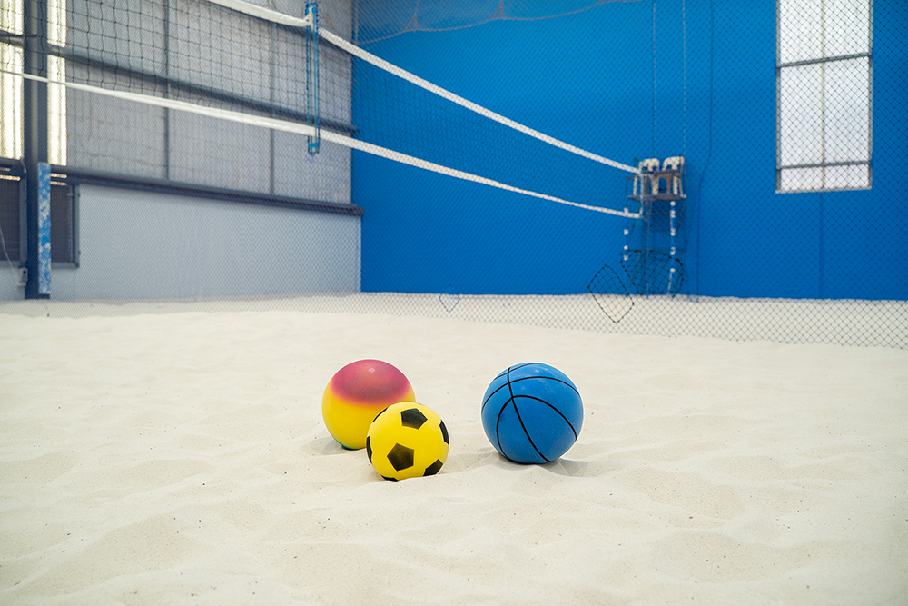 Colourful sports balls on a sand court inside an indoor sports facility with volleyball net.
