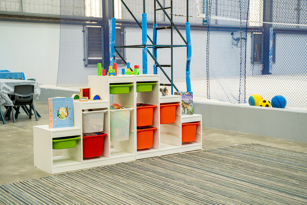 White storage shelving with colourful bins, blocks, and books in an OSHC play area, with sports balls nearby.