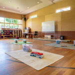 Spacious OSHC hall with timber floors, activity mats with toys, storage shelving, whiteboard, and high windows.