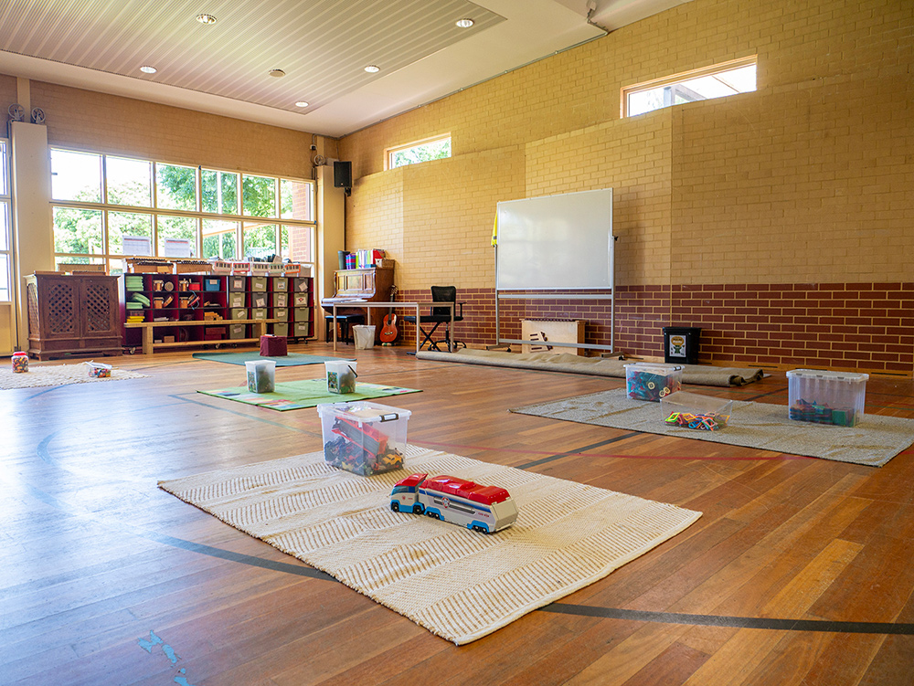 Spacious OSHC hall with timber floors, activity mats with toys, storage shelving, whiteboard, and high windows.