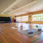 Large OSHC hall with polished timber floors, activity mats, colourful banners, stage area, and floor-to-ceiling windows.