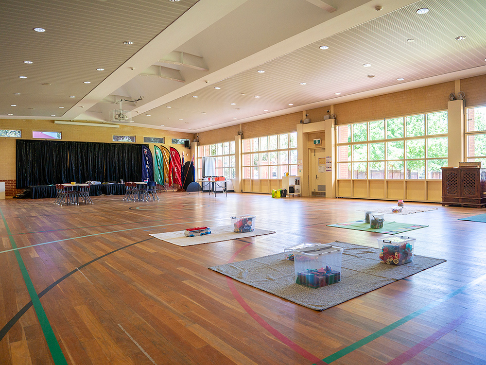 Large OSHC hall with polished timber floors, activity mats, colourful banners, stage area, and floor-to-ceiling windows.