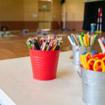 Colourful pencils, markers, and scissors in metal buckets on a table in an OSHC craft area.