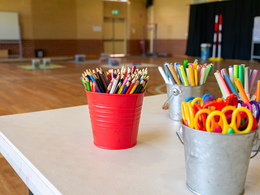 Colourful pencils, markers, and scissors in metal buckets on a table in an OSHC craft area.