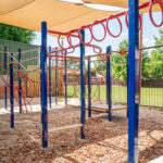 Shaded outdoor playground with blue and red climbing frame equipment on sand, with green oval beyond the fence.
