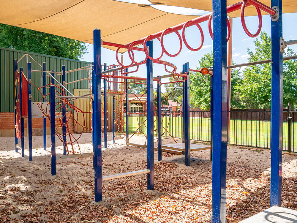 Shaded outdoor playground with blue and red climbing frame equipment on sand, with green oval beyond the fence.