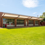 Exterior of a brick OSHC building with large windows, covered walkway, and manicured green lawn.