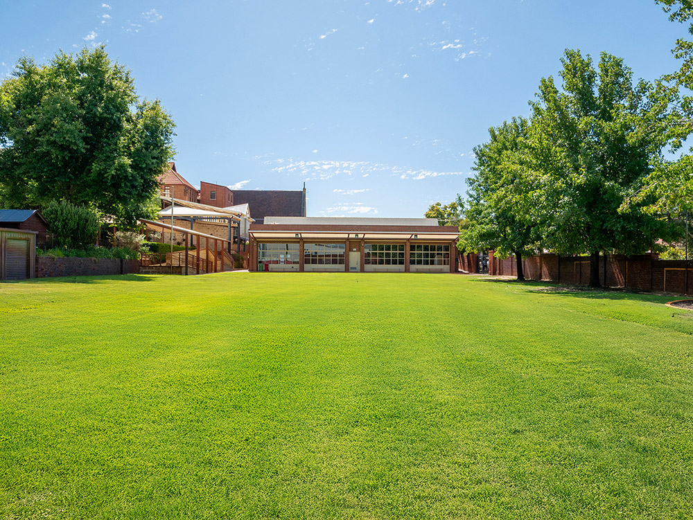 Spacious green oval with mature trees, OSHC building, and covered outdoor area in the background.
